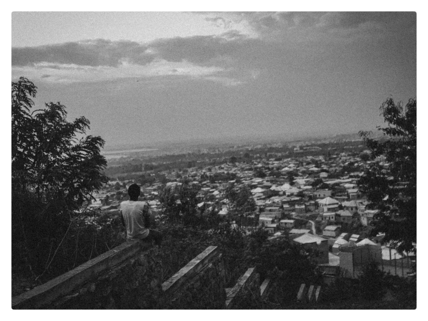Monochrome shot of a person sitting on a ledge overlooking a dense cityscape surrounded by trees, captured in a natural outdoor setting.