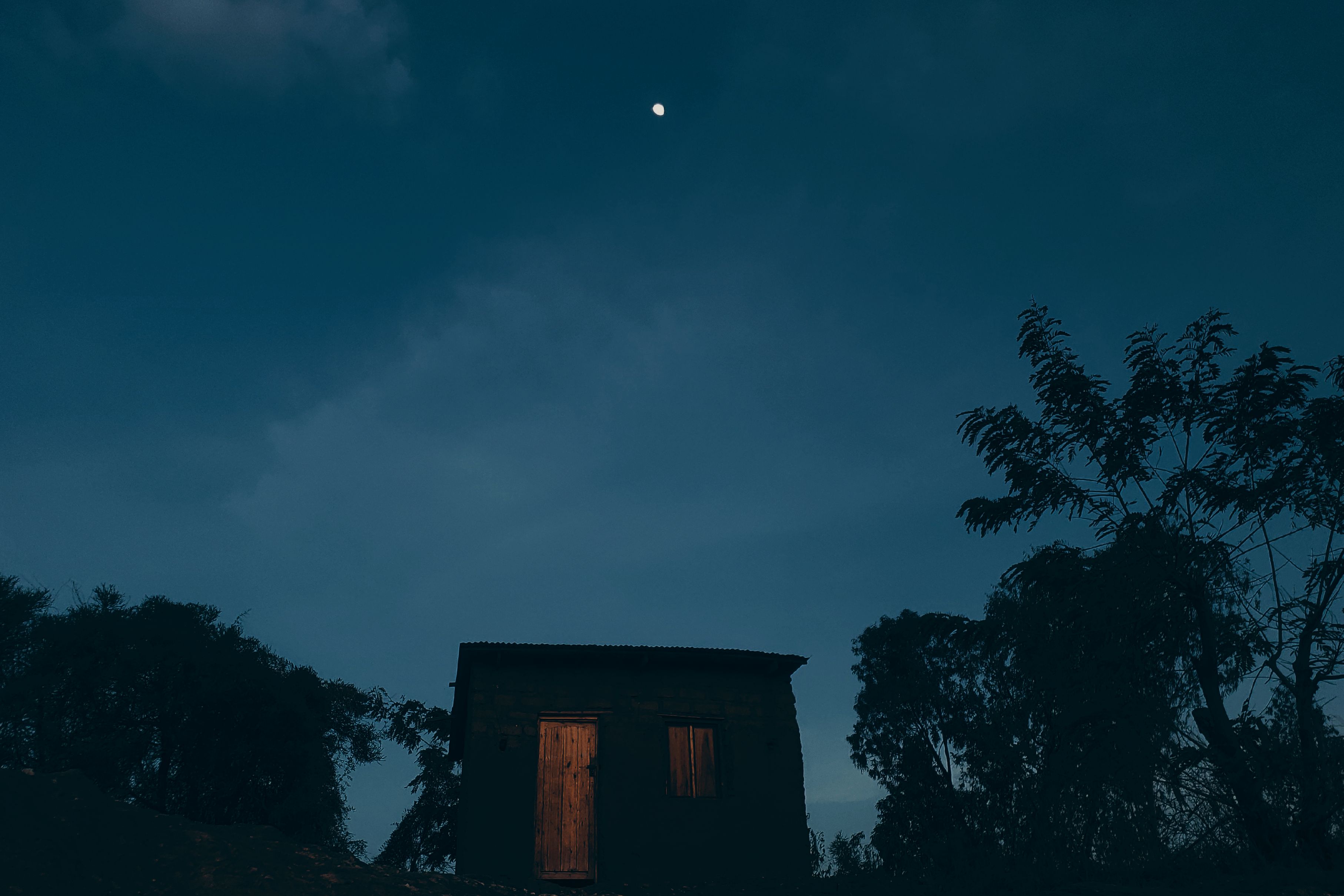 A small, dark house at night, with a glowing wooden door and visible window, set against a backdrop of trees and a blue sky.
