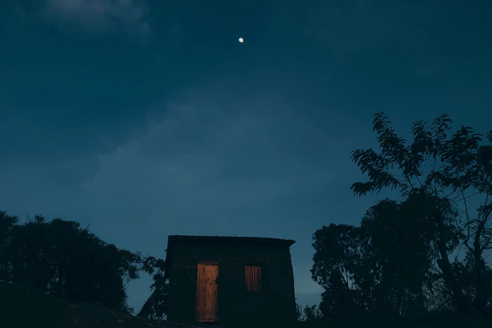 A small, dark house at night, with a glowing wooden door and visible window, set against a backdrop of trees and a blue sky.