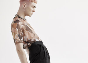Profile shot of a young adult with short pink hair, wearing a patterned top and black pants, against a seamless white background.