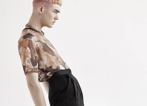Profile shot of a young adult with short pink hair, wearing a patterned top and black pants, against a seamless white background.