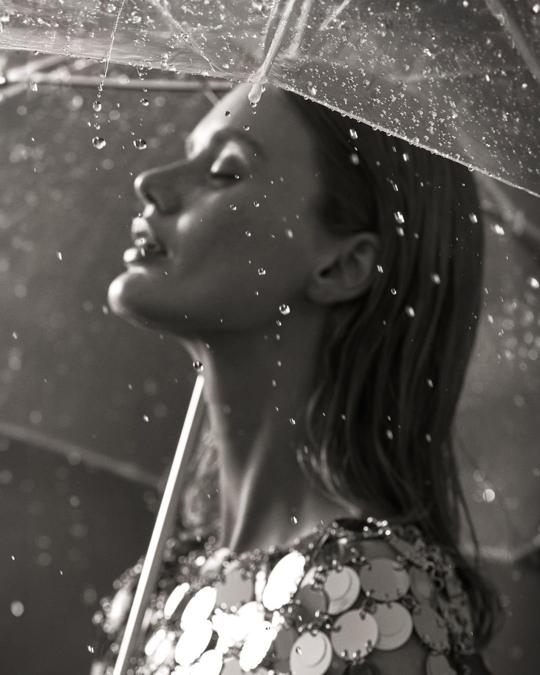 Monochrome portrait of a woman with her eyes closed under a clear umbrella, capturing falling raindrops and wearing a sequin dress.