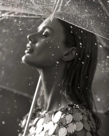 Monochrome portrait of a woman with her eyes closed under a clear umbrella, capturing falling raindrops and wearing a sequin dress.