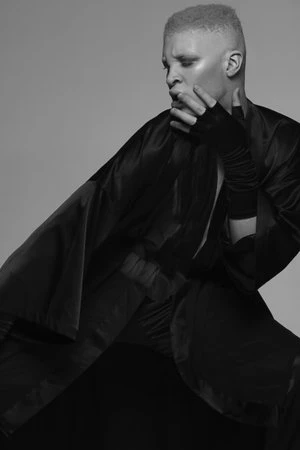 Black and white portrait of androgynous person wearing dramatic black satin cape, gloves, and pants, posing with hand to face in a studio.