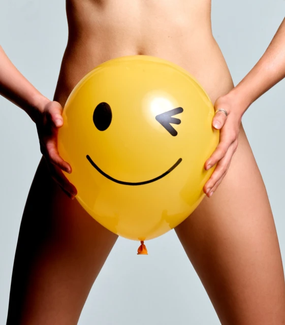 Unidentified person holds a yellow balloon with a winking smiley face. Arms are visible with a plain background.