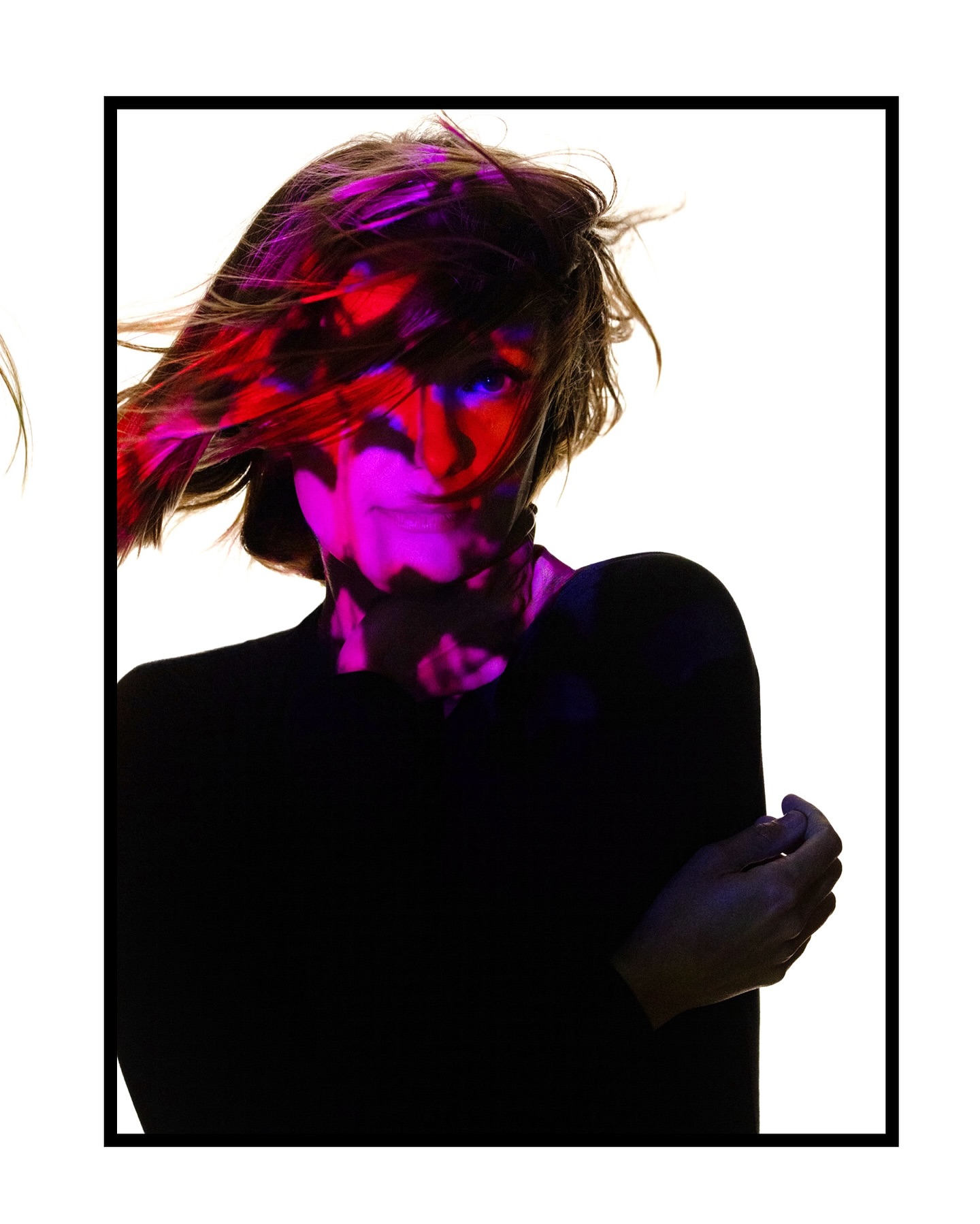 A woman is shown in a close-up indoor portrait with red and purple lighting on her face, against a bright white background.