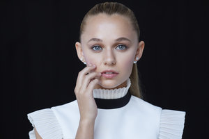 Portrait of a young, fair-skinned female with blond hair in a ponytail, wearing a white top, posing in front of a dark background.