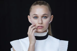 Portrait of a young, fair-skinned female with blond hair in a ponytail, wearing a white top, posing in front of a dark background.