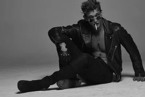 Man wearing a leather jacket and sunglasses holding a cigarette while seated on a studio floor.