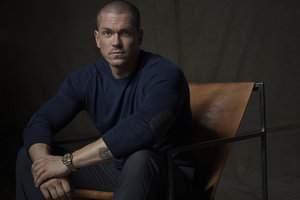 Man with short hair seated in a leather chair wearing a dark blue sweater and dark trousers photographed by Frankie Batista