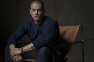 Man with short hair seated in a leather chair wearing a dark blue sweater and dark trousers photographed by Frankie Batista