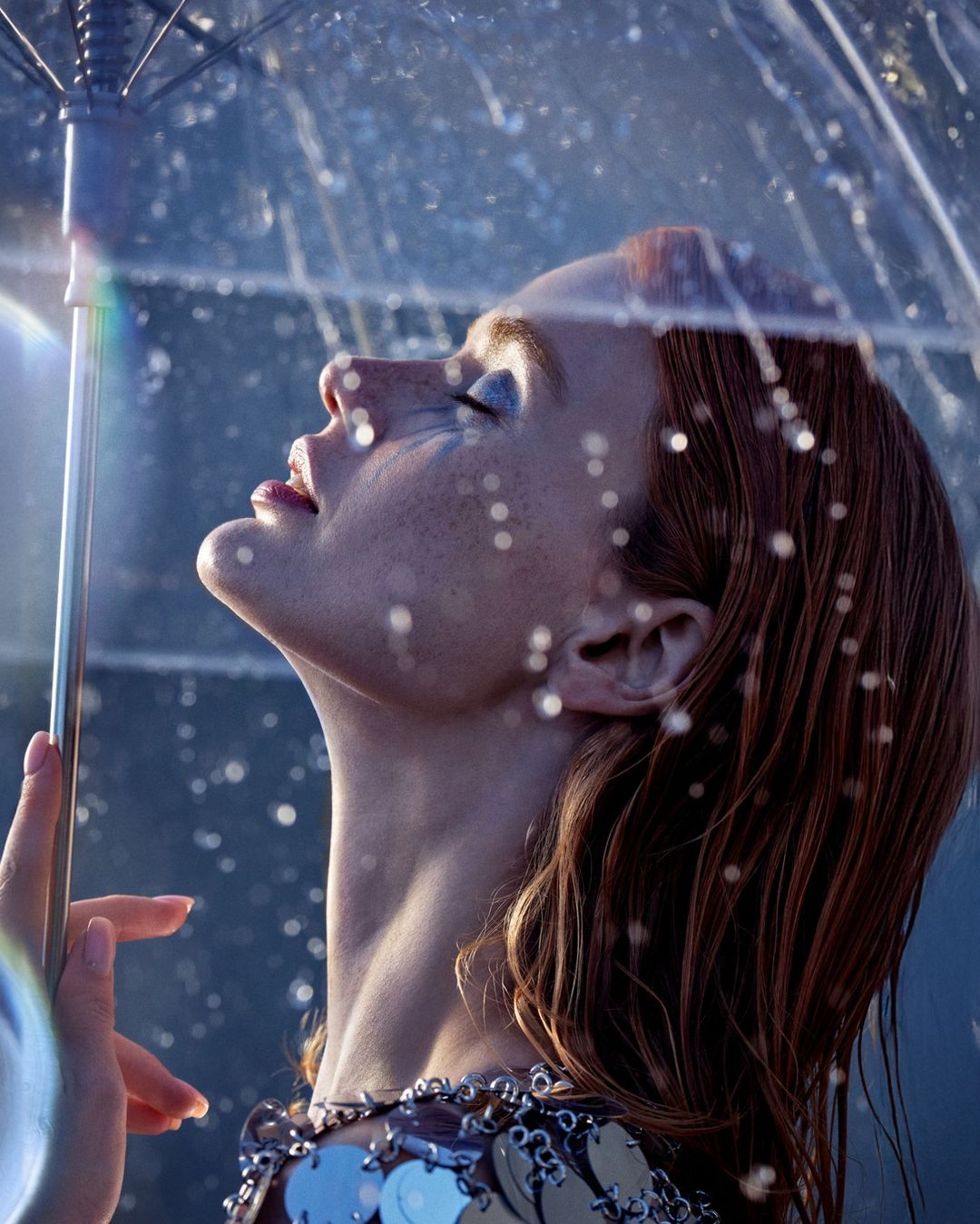 Close-up of a woman with red hair looking up with a transparent umbrella, wearing a silver dress with sequin details.