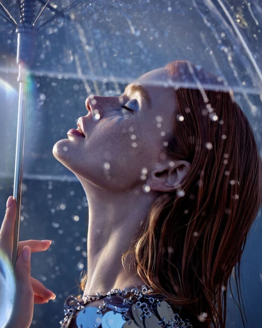 Close-up of a woman with red hair looking up with a transparent umbrella, wearing a silver dress with sequin details.