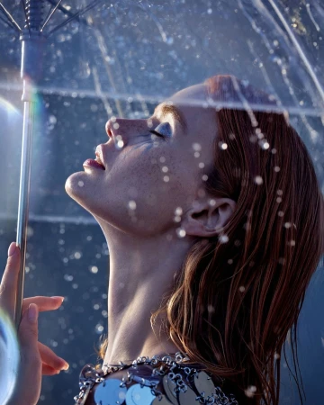 Close-up of a woman with red hair looking up with a transparent umbrella, wearing a silver dress with sequin details.