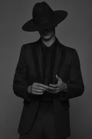 Man wearing a wide-brimmed black hat and dark suit standing against a grey background in a portrait photograph by Frankie Batista.