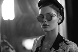 Monochrome portrait of a woman wearing round sunglasses and a dark cap by photographer Frankie Batista.