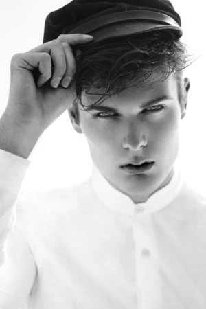 Monochrome portrait of a male model wearing a white shirt and cap, his hand gently touching the cap's brim against a light background.