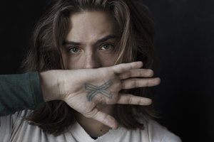 Close-up portrait of a man with long hair holding his hand with an X tattoo in front of his face by Frankie Batista