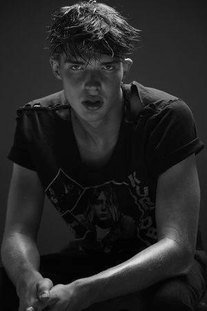 Black and white portrait of a young adult man with wet, styled hair, wearing a graphic tee-shirt in a studio setting.