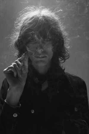 Portrait of a man with long hair touching his cheek surrounded by smoke in monochrome lighting by Frankie Batista.