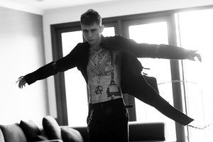 Monochrome portrait of a young man with extensive torso tattoos wearing a dark open jacket and chain necklace