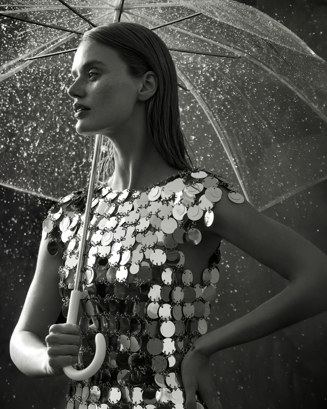 Monochrome portrait of a woman with wet hair under a clear umbrella, wearing a silver sequin dress, looking off to the side.