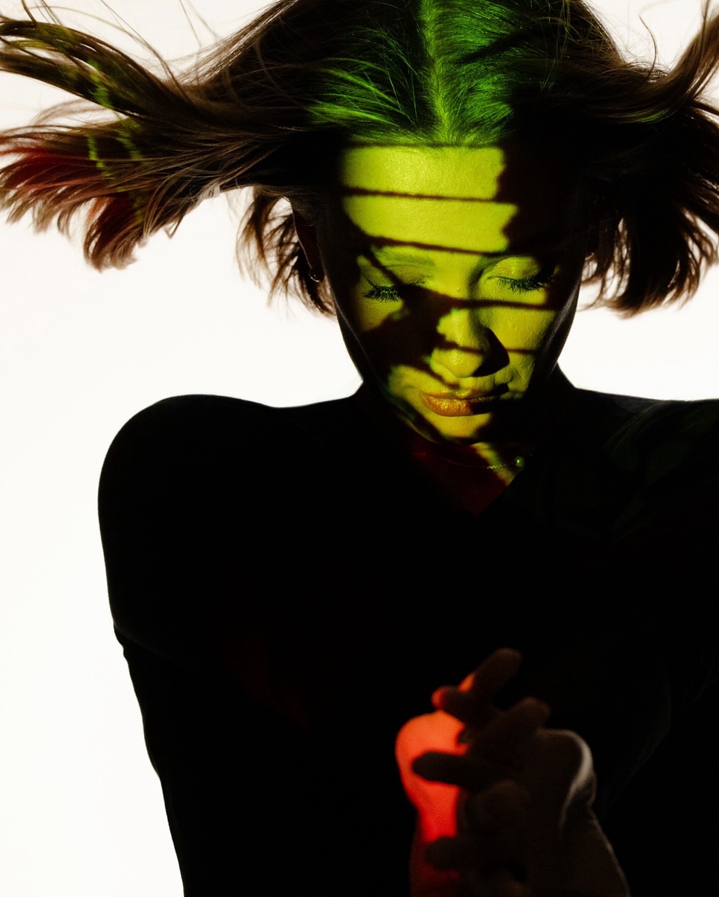 Dramatic portrait of a woman with dark hair, bathed in green and red light with a white background. Close-up captures intense mood.