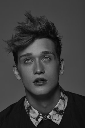 Monochrome portrait of a young adult male with styled hair, wearing a floral-collared shirt, looking upward against a gray background.