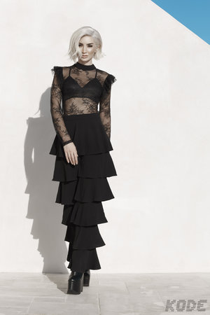 A woman with short white hair wears a black lace top and a layered skirt, standing in front of a white wall with a blue accent.
