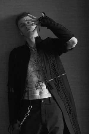 Black and white studio portrait of a slender man with tattoos, wearing a black jacket and pants, covering his face with his hand.
