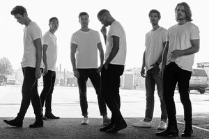 Six men stand in a parking lot wearing white t-shirts and dark pants in a monochromatic outdoor shot, with buildings in the background.