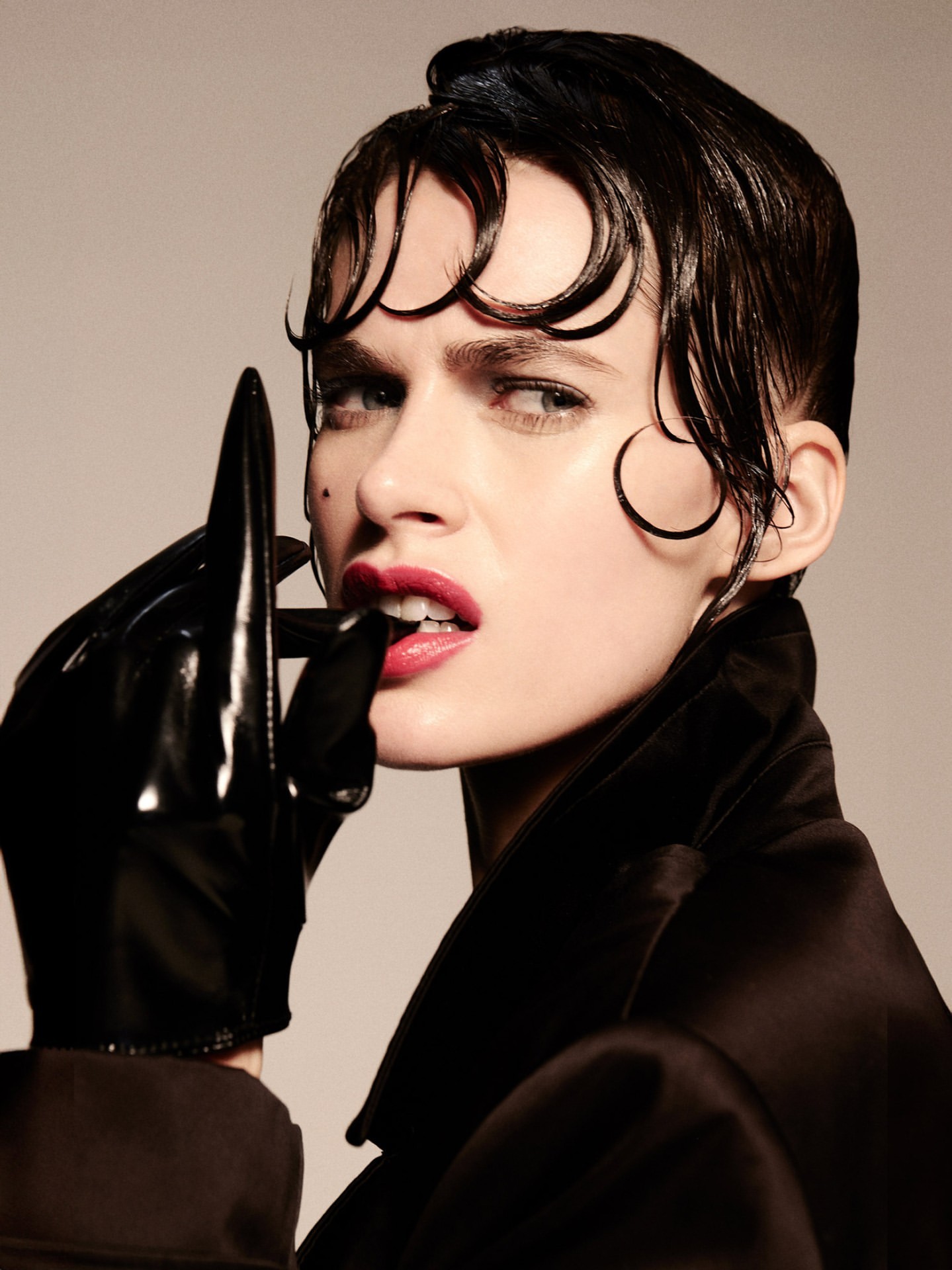 Close-up portrait of a woman with finger in mouth, wearing shiny black gloves and a dark jacket against a light background.