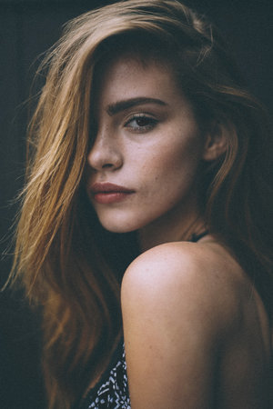 Portrait of a fair-skinned adult female with brown eyes and long auburn hair, looking over her shoulder in a dimly lit environment.