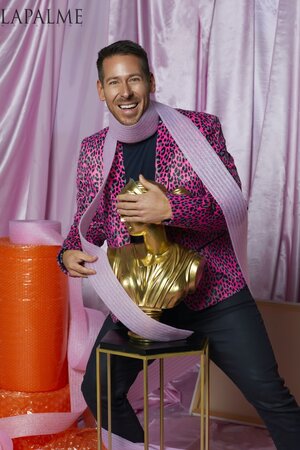 A man in a pink leopard print blazer smiles while holding a gold bust wrapped in pink foam, set against a pink draped background.