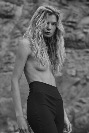 Monochrome portrait of a blonde woman with wavy hair wearing black pants posing outdoors against a rock background.
