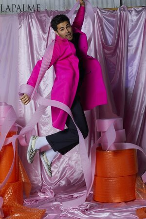Man jumping in a pink coat surrounded by pink bubble wrap rolls in a surreal studio environment with a soft, dreamy aesthetic.