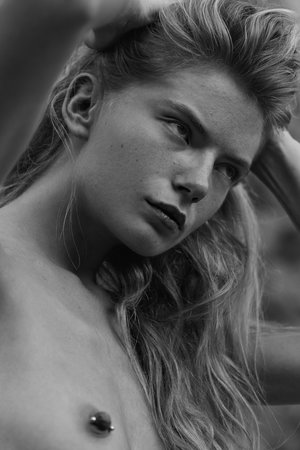 Black and white portrait of a young adult woman with long wavy hair and a nipple piercing, hands behind her head, looking to the side.