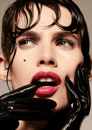 Glamorous beauty portrait of a woman with slicked-back hair, red lips, and shiny black gloves, set against a soft gray background.