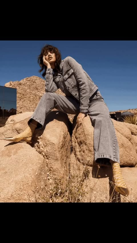 Woman in denim jacket and jeans sits on desert rock, wearing tan boots against a clear blue sky in a fashion shot.