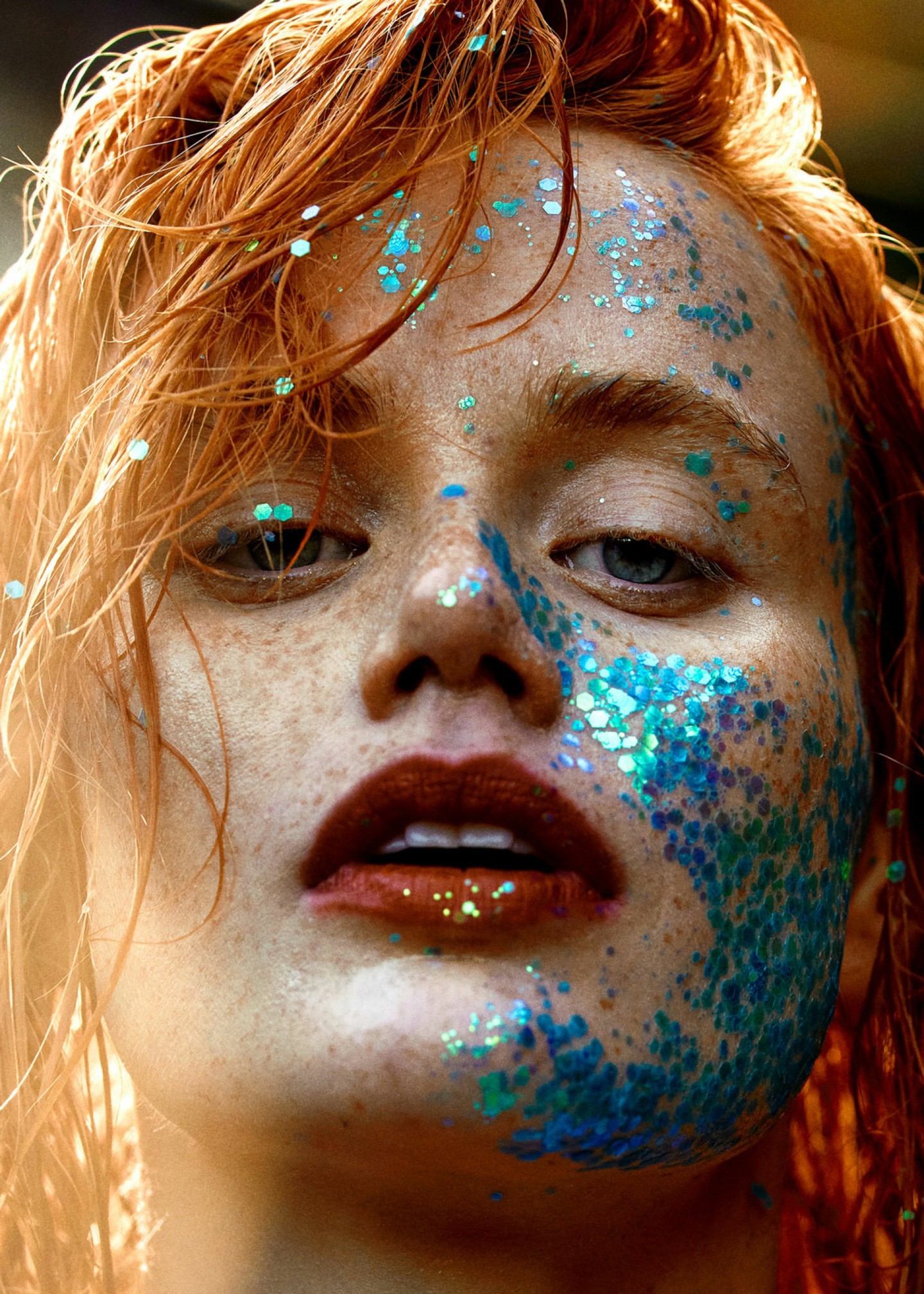 Close-up portrait of a red-haired woman with freckles and blue glitter makeup on her face, highlighting her eyes and lips.
