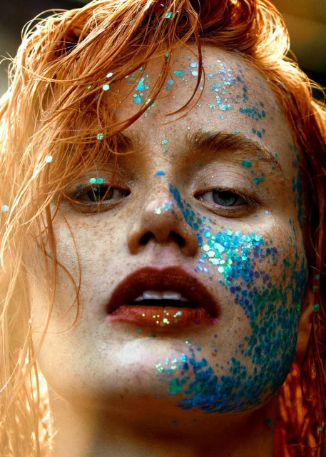 Close-up portrait of a red-haired woman with freckles and blue glitter makeup on her face, highlighting her eyes and lips.