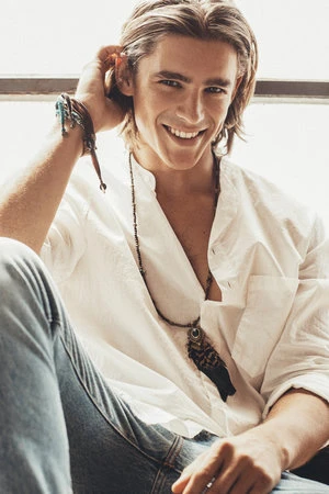 Portrait of a smiling young man with long blonde hair wearing a white shirt and beaded necklaces by photographer Frankie Batista.