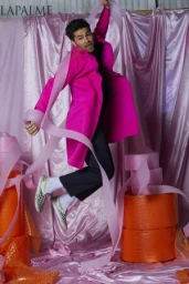 Man in a bright pink coat and dark trousers jumping mid-air amidst pink satin fabric and orange bubble wrap rolls