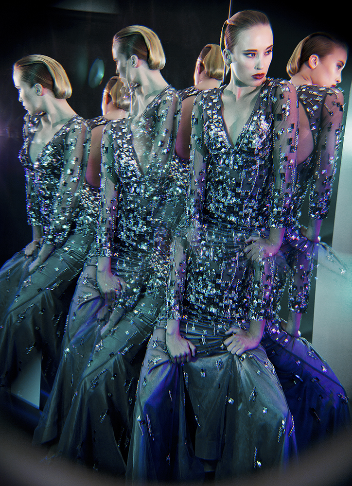 A woman wearing a long, shimmery gown poses among multiple reflections, creating a surreal and glittering visual effect.