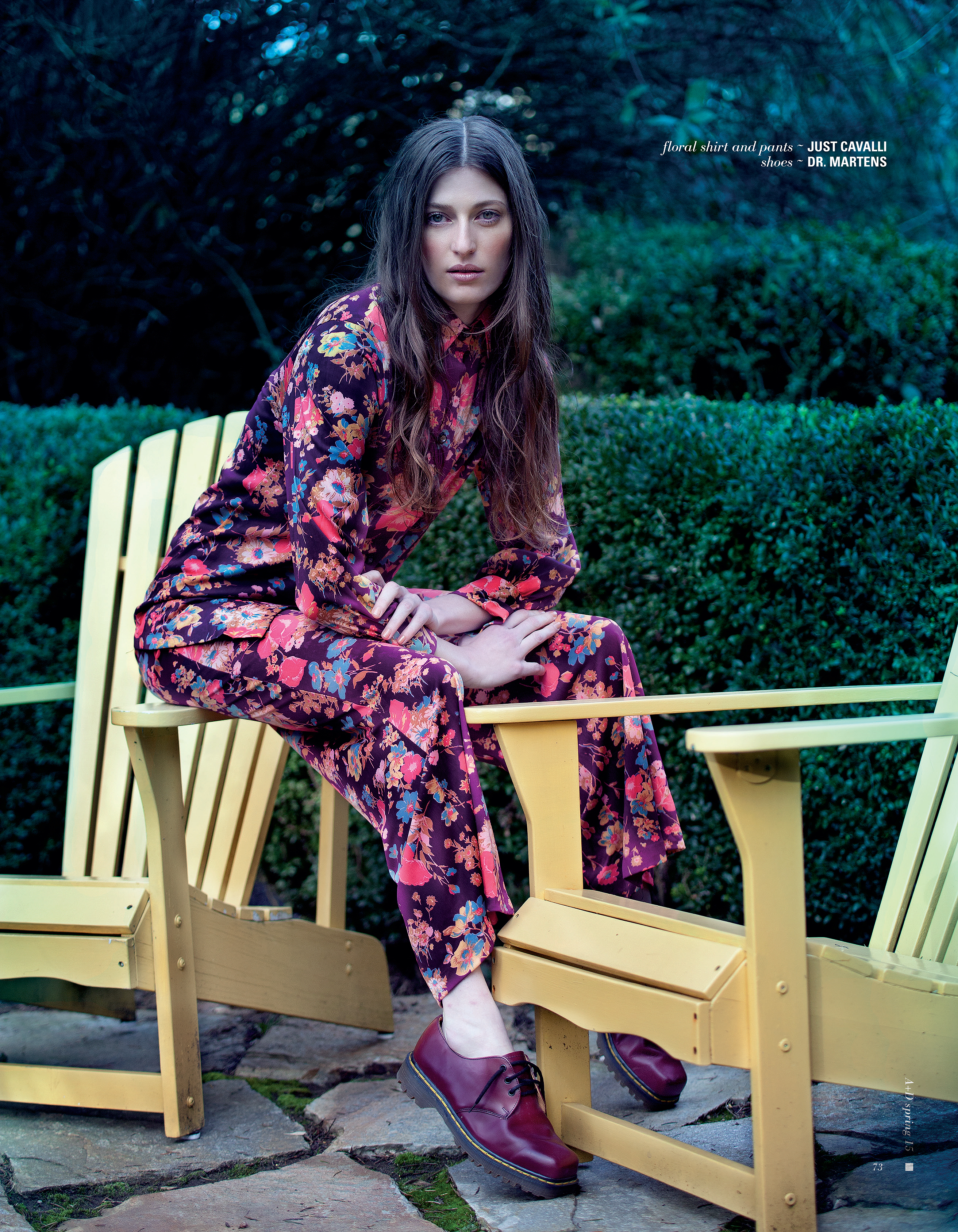 An adult female model sits on a yellow chair in a floral print suit with maroon Dr. Martens, outdoors with a green hedge background.