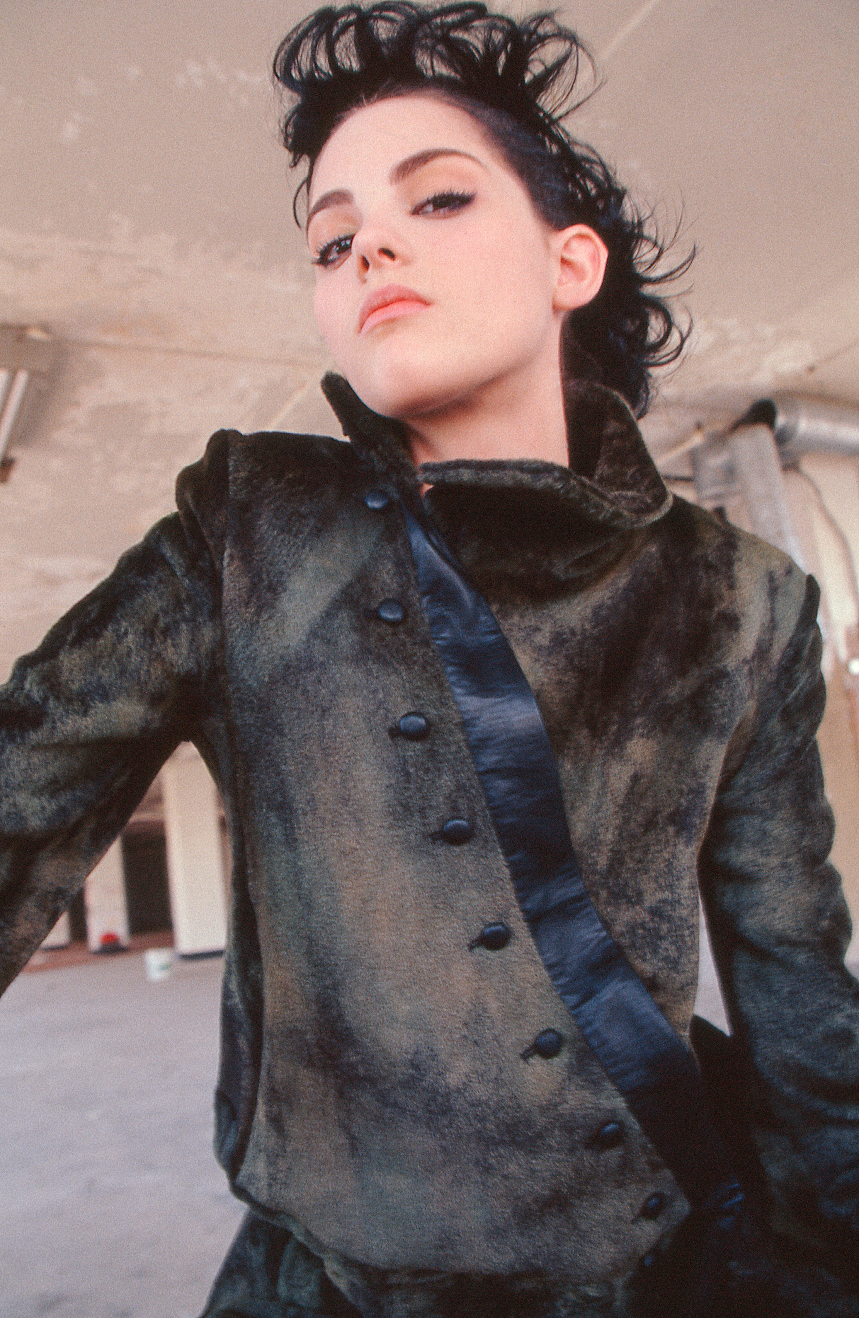 Stylish woman in a vintage-inspired dark fur coat poses indoors, looking down from a low angle, with natural light and a minimalist backdrop.