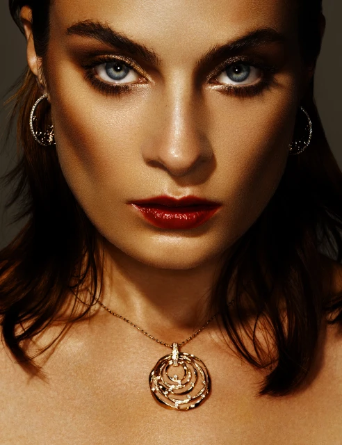 Close-up portrait of a woman with dark hair, red lipstick, and blue eyes, wearing gold necklace and earrings against a grey background.