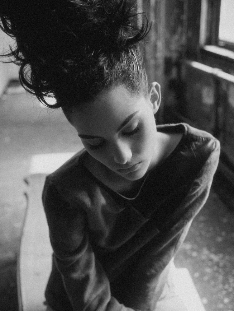 Black and white portrait of a woman with dark hair styled up, wearing a long-sleeve shirt, seated in a room with natural light.