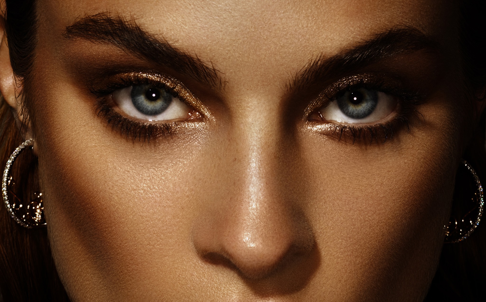 Close-up of a woman's face featuring her eyes with gold eyeshadow and hoop earrings, set against a dark background.