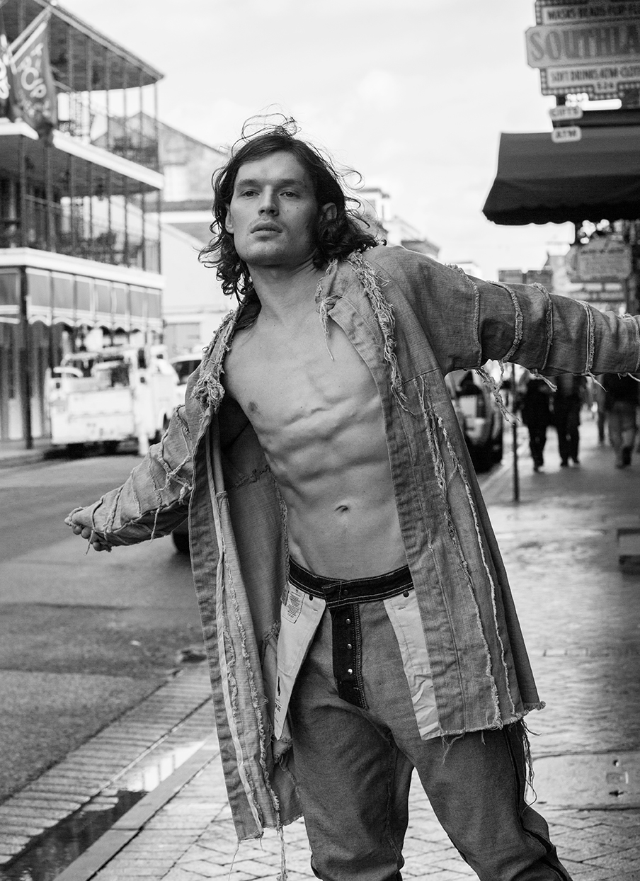 Black and white shot of a shirtless man in a jacket and jeans in an outdoor urban setting with buildings and signage in the background.